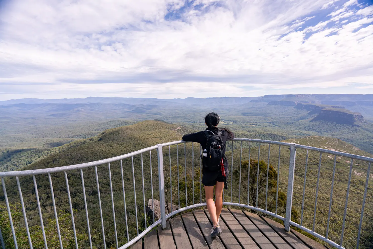 Woman looking out at a 360 degree view at the top of a mountain. Higher than the tree tops. Mountains can bee seen far in the distance.