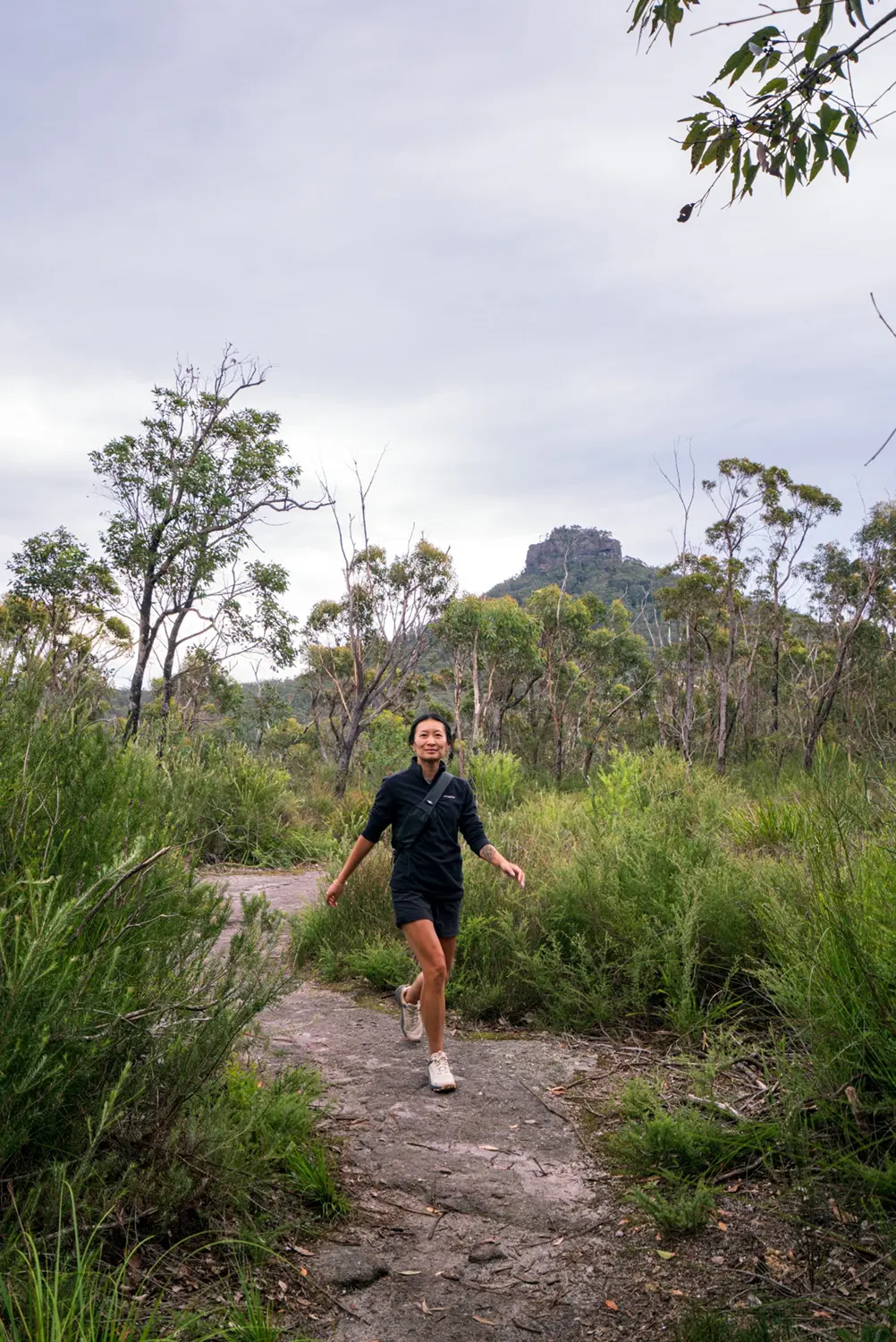 Woman walking towards the camera on a trail. Forest around her and a mountain in the distance behind her.
