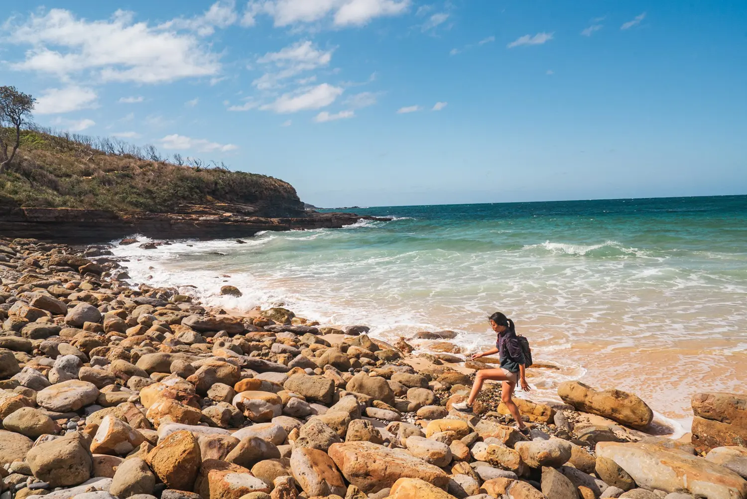 Woman talking over large stones and rocks overing the entire beach. Water and whitewash to the right and highland in the background.