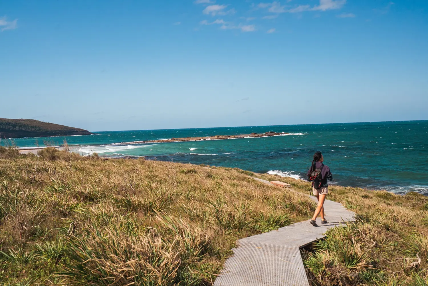 Woman walking on a coastal trail on a steel mesh walkway that winds around the corner. Blue green ocean to the right and a beach far in the background